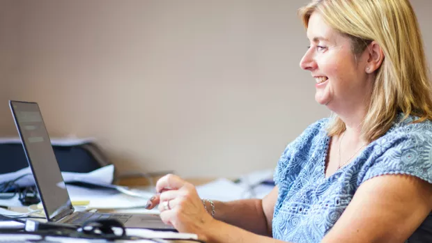 Photo: a woman sat at a desk looking at the MS forum on a laptop she is smiling