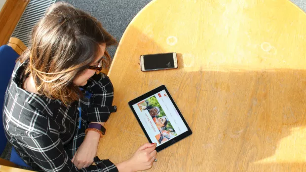 Photo shows a woman sitting at a table with an iPad and smartphone.