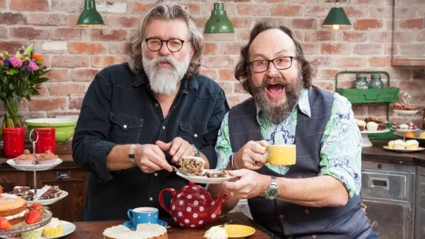 The Hairy Bikers in a kitchen, holding cake and a cup of tea and smiling