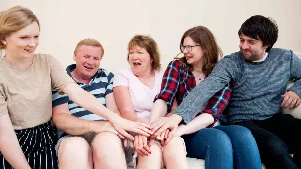 Photo shows a family of two parents and three grown-up children sitting together, laughing. 