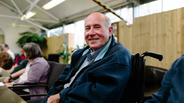 Photo shows a man in a wheelchair smiling at the camera, with his friends behind him. 