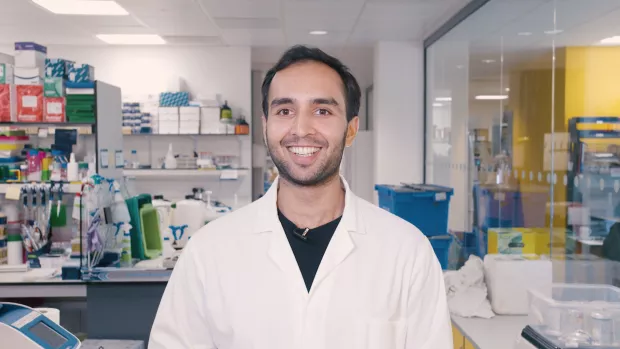 Image shows Amar in a lab coat smiling at camera against background of science lab