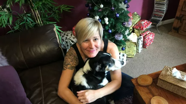 Caz cuddles her dog. Behind them is a Christms tree surrounded by presents.