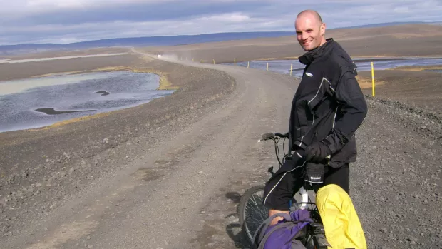 Ewan stands with his bicycle in front of a long road that winds far into the horizon