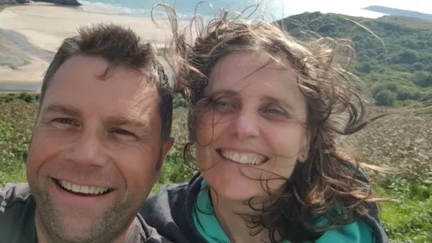 Florence with her husband John, smiling with a beach pictured behind them.  