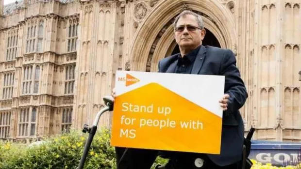 Photo of man in chair outside Westminster with placard that says stand up for MS