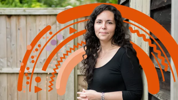 Woman with curly hair holding a mug stands in front of her garden fence with an orange rainbow behind her 