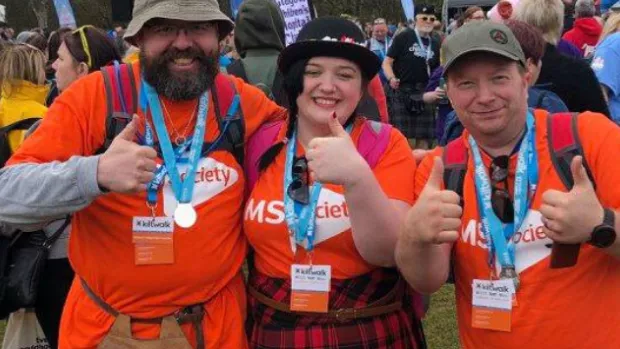 Two men and a woman take a moment out from the crowd, all wearing hats, MS Society shirts and kits smile with thumbs up at the camera sporting their medals for completing the Kiltwalk