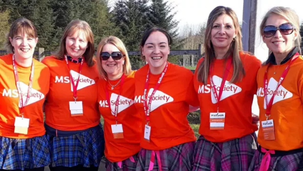 Seven women wearing MS Society t-shirts and colourful kilts smile to camera on a bright sunny day 