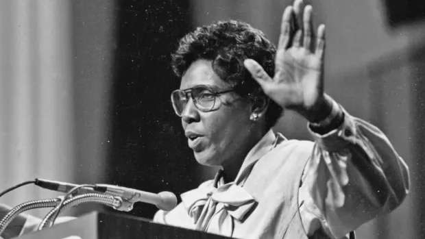 Black and white photo of Barbara Jordan speaking at a podium with her left hand raised palm up