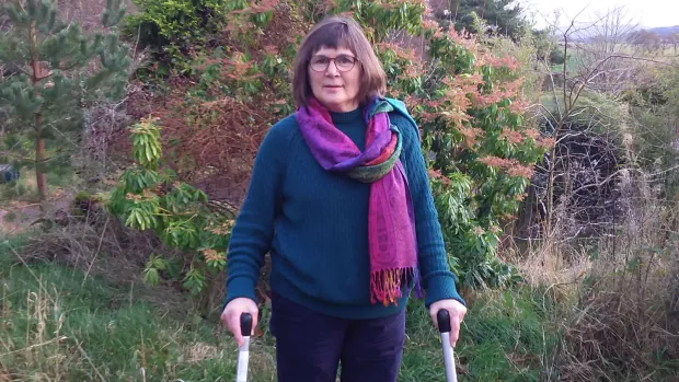 Nina stands outside with the aid of two walking sticks. She has brown hair, glasses and is wearing a green jumper.