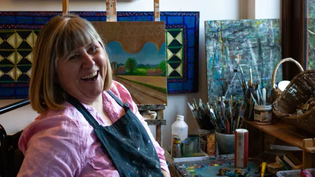 Jenny sits surrounded by art materials, wearing a paint-splattered apron and with a big smile