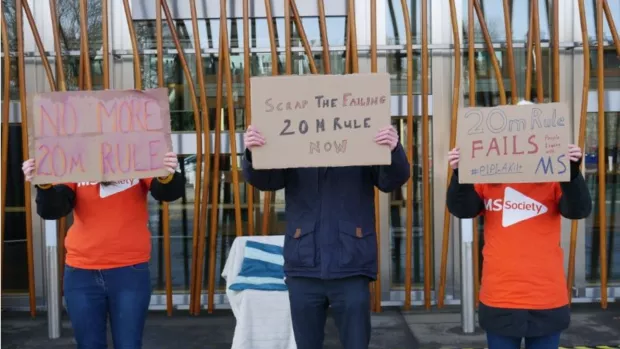 Three people hold us signs about scapping the 20 metre rule, outside the Scottish Parliament.