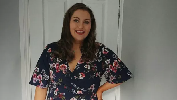 Colleen stands in a room smiling and wearing a floral dress.