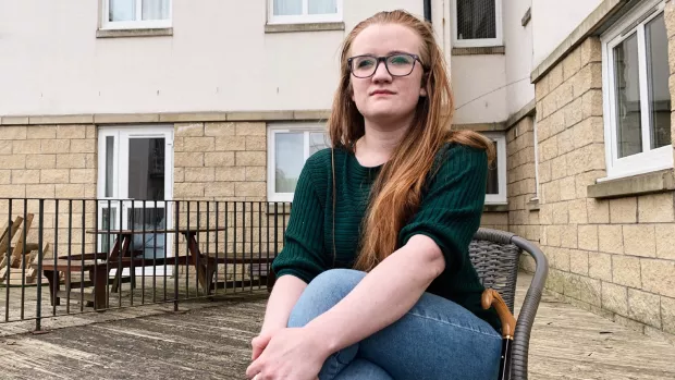 Fiona sits on a chair outside a block of flats. She has long red hair and is wearing glasses.