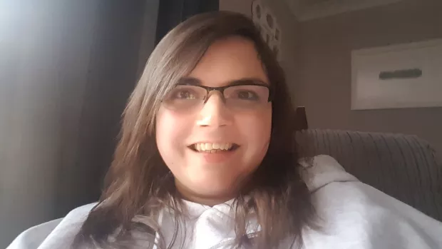 Kirsty sits at home. She is wearing glasses, has long brown hair and is smiling. 