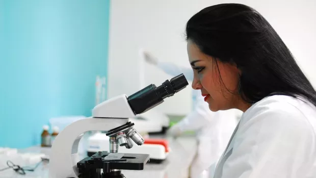 A scientist looks into a microscope in a lab. She has long black hair.