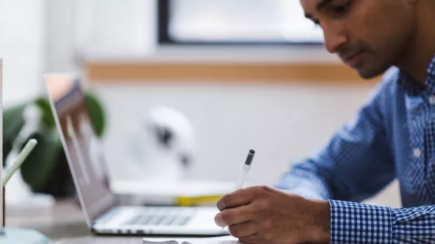 A man in a blue and white checked shirt is writing in a note page and has a laptop open on his desk