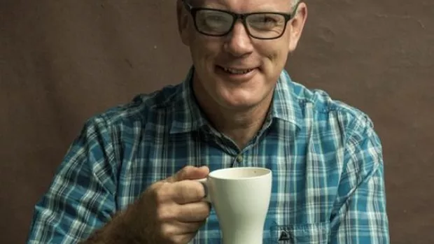 A man in a green checked shirt is smiling and holding up a white mug