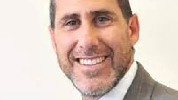 Dr Eli Silber, a headshot of a man in a grey checked suit with peppery stubble and short cropped dark hair