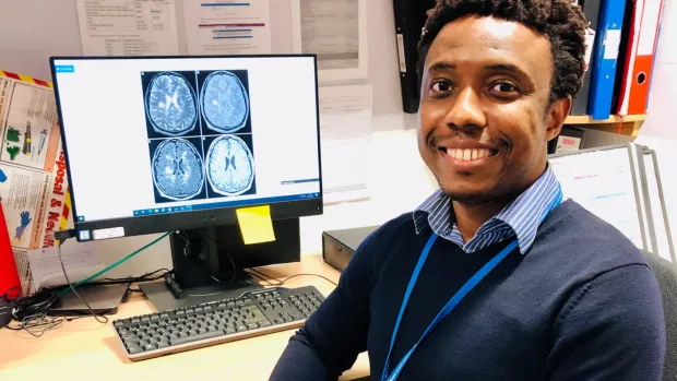 This image shows a man in a shirt and jumper smiling at the camera in front of a computer on a desk showing four MRI brain images 