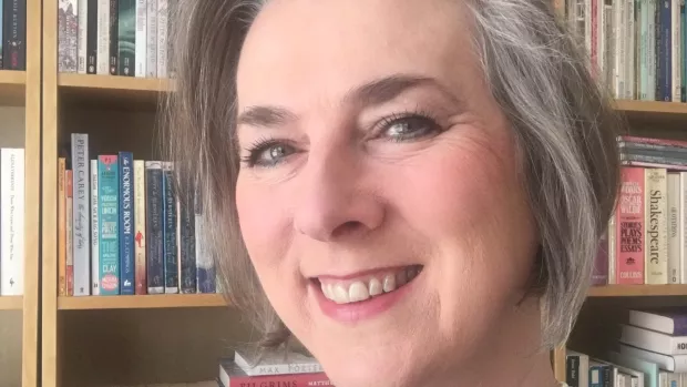 close up of a smiling woman with grey hair stood in front of a bookcase. 