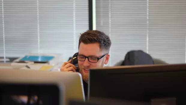 A man with glasses and a beard talks on a telphone in front of a computer monitor.