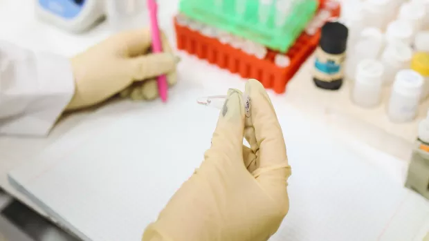 two hands in latex gloves holding a test tube and writing in a book