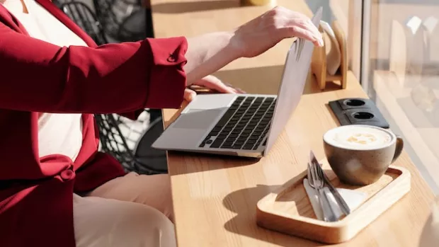 A woman in a red jacket is sat in a wheelchair at a coffee shop window with an open laptop and a cup of coffee