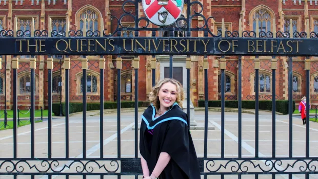 A photo of Anna in her graduation gown in front of The Queen's University of Belfast gates