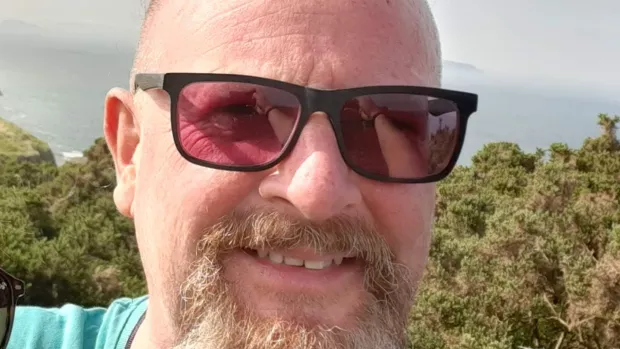 A photo shows Iain smiling and wearing sunglasses outside in the sun