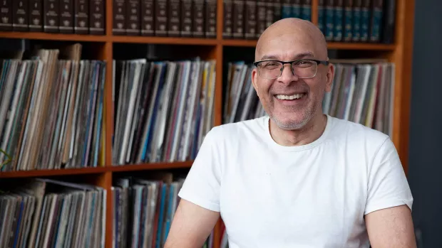 Image of Robby sitting in his flat, smiling. Behind him are shelves full of his record collection.