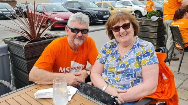 Ann is sitting outside at an MS Society event. She's wearing sunglasses and a bright t-shirt with flowers on.