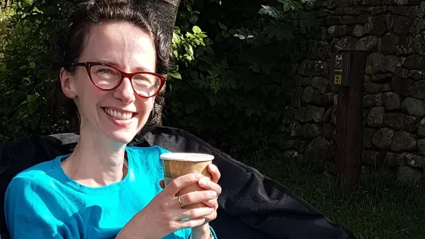 Jo sits outside smiling in the sun and holding a cup.