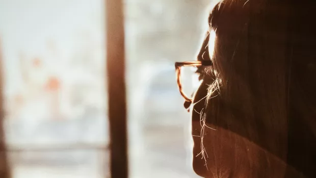 The photo shows a woman wearing glasses looking out of a window. 