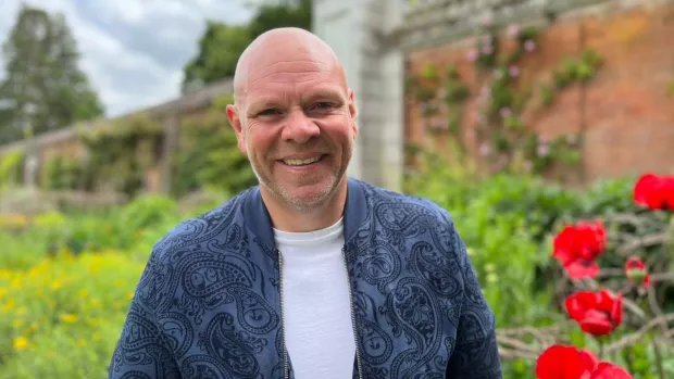 Tom Kerridge stands outside next to some red poppies smiling at the camera