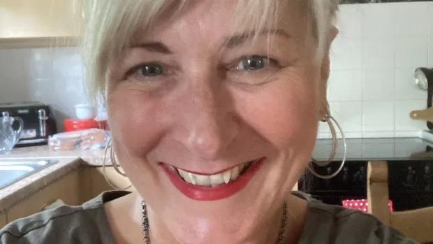 A selfie of a smiling woman wearing red lipstick and gold hoop earrings, she's stood in her kitchen