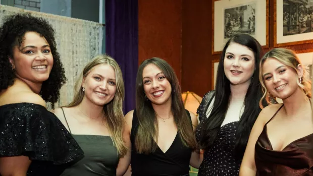 Five young people stand shoulder to shoulder, arms around each other's backs, they're smiling and in party dresses