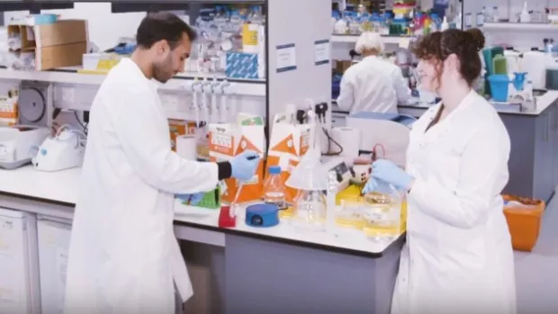View of a lab with three scientists in white labcoats, a man and a woman in conversation at a bench, and a man carrying a large beaker of yellow liquid