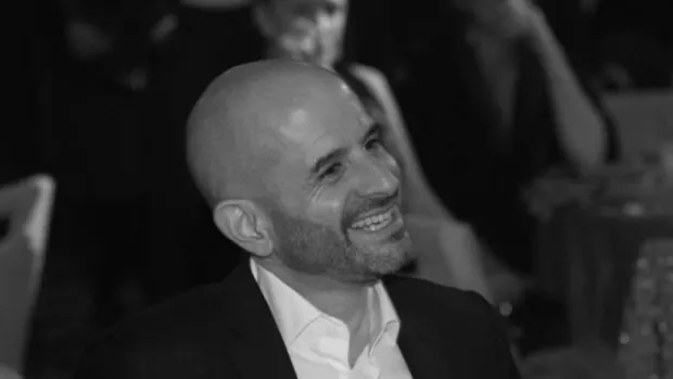 The photo shows David sitting at a table, smiling and dressed in a smart shirt and jacket. 