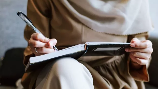 Image shows a person's torso and lab. The person is wearing beige clothing, has a notepad balanced on one knee, and writing with a pen. 