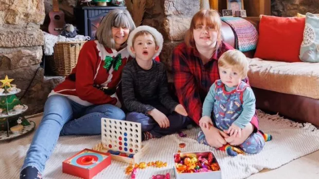 A family are sat on the floor in front of a stone fireplace, one person wears a red Christmas jumper and sits with a child wearing a Santa hat, around them are games and chocolates and cakes on a stand shaped like a Christmas tree. 
