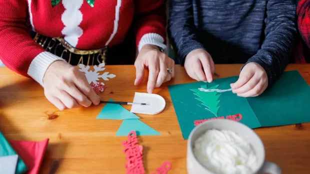 A child and adult sit at a table making Christmas cards with green and red card. The adult wears a festive jumper liken an elf's outfit which is red with white trim.