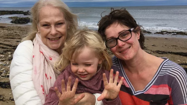 Katherine and her daughter Ellie stand on a beach holding Ellie's young daughter. all are smiling.