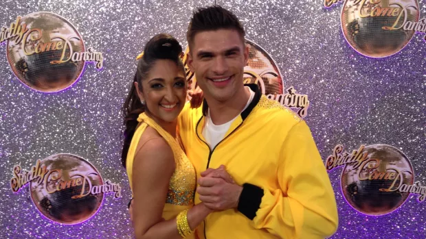 Trishna and Aljaz are wearing yellow and posing together in front of a Strictly Come Dancing backdrop.