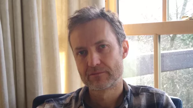 A man with grey hair and stubble sits in front of a window through which streams light. He's wearing a plaid shirt. 