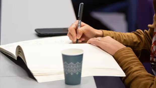On the right is the torso of a person in a camel coloured long sleeve shirt and MS Society orange and white lanyard. They're sat at a grey desk with a blue disposable coffee cup and mobile phone. They're writing in an A4 journal. 