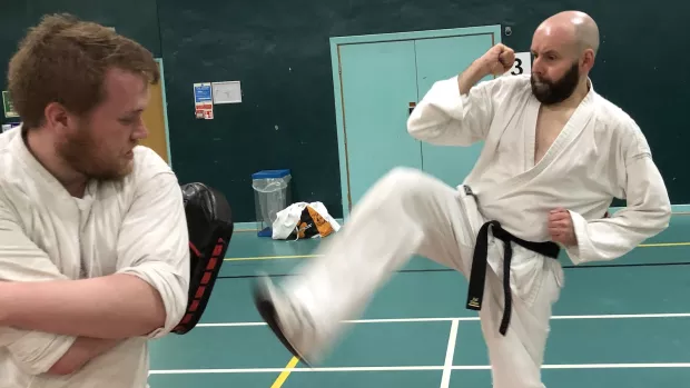 two men in a green gymnasium practicing karate. On the right Scott, in white karate Dogi and black belt, kiks at a fellow club member who is defending with a pad held up under is right arm