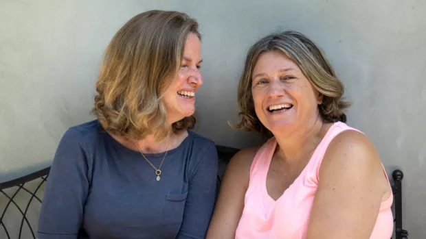 Nina and Milla are sitting side-by-side on a bench, smiling at each other and the camera.