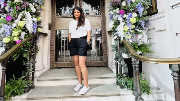 A woman in black shorts and a white shirt stnads on marble steps in front of a large door garlanded with flowers